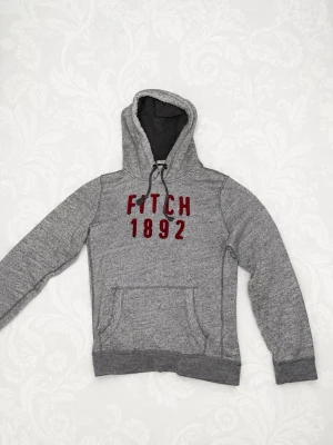 Grå hoodie Abercrombie & Fitch - Snygg grå hoodie från Abercrombie & Fitch med röd text 'FITCH 1892' på bröstet. Tröjan har magficka, dragsko i huvan och mjukt fleecefoder på insidan. Perfekt för chill dagar eller när du vill ha en avslappnad stil.        Ända anledningen att jag säljer är att den är lite för kort för mig, jag själv är 189 så passar nog fint under det👍