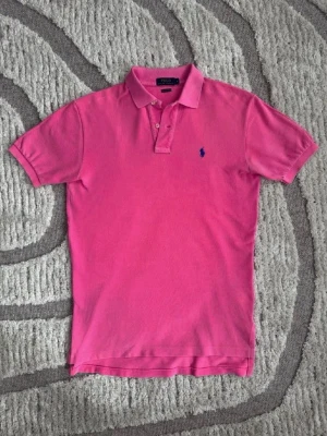 Rosa pikétröja från Polo Ralph Lauren - Säljer en klassisk rosa pikétröja från Polo Ralph Lauren med blå logga broderad på bröstet. Tröjan har korta ärmar, krage och knappar vid halsen. Perfekt för en avslappnad men ändå stilren look.