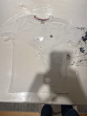 Vit t-shirt från Moncler - Säljer en stilren vit t-shirt från Moncler med klassisk rund hals och korta ärmar. T-shirten har Monclers ikoniska logga broderad på bröstet och är tillverkad i mjuk bomull. Perfekt till jeans eller shorts för en clean look.
