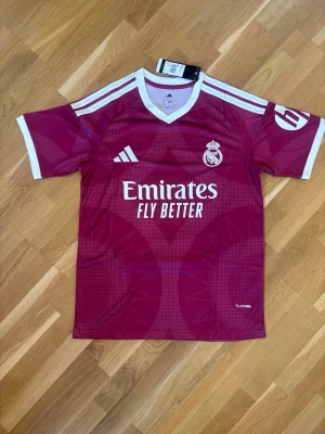 Real Madrid Mbappé matchtröja vinröd - Snygg vinröd Real Madrid matchtröja från Adidas med vita detaljer och tryck. Tröjan har klubbmärke, Adidas-logga och sponsortryck framtill samt Mbappé och nummer 10 på ryggen. Tillverkad i lätt och ventilerande material, perfekt för fotboll eller träning.
