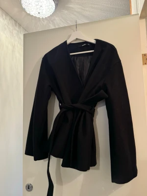 Svart kappa med knytskärp - Stilren svart kappa med knytskärp i midjan och rak passform. Kappan har en öppen design utan synliga knappar och är helfodrad för extra komfort. Perfekt för dig som gillar minimalistisk och tidlös stil.