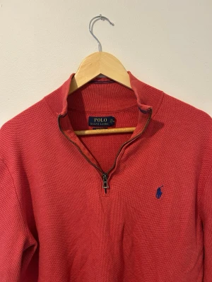 Röd half zip tröja från Polo Ralph Lauren - Snygg röd tröja från Polo Ralph Lauren i mjuk pima bomull. Klassisk modell med half zip och blå broderad logga på bröstet. Perfekt till jeans eller chinos för en clean och stilren look.