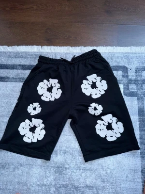 Svarta shorts med vita blommor - Svarta shorts med elastisk midja och vita blommönster på både fram- och baksida. De har en avslappnad passform och är perfekta för sommaren. Shortsen har fickor på sidorna och en bakficka. Snygg och trendig design för en chill look.