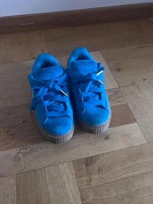 Blåa Puma Fenty sneakers - Snygga blåa Puma Fenty sneakers med tjock sula och gula detaljer på sidan. Skorna har breda skosnören med guldfärgade metalldetaljer och är tillverkade i mockaliknande material. Perfekta för dig som vill sticka ut med färg på fötterna.