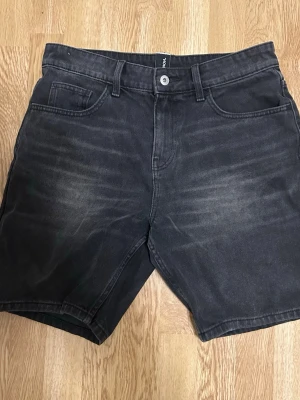 Svarta denimshorts med normal passform - Säljer ett par svarta denimshorts med klassisk femficksdesign och bälteshällor. Shortsen har en knapp och dragkedja framtill samt en något tvättad look för en avslappnad stil. Perfekta till sommaren och lätta att matcha med olika outfits.