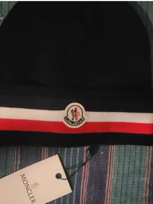 Svart Moncler mössa med ränder - Snygg svart mössa från Moncler med klassiska vita och röda ränder samt Moncler-logga framtill. Mössan har uppvikt kant och är tillverkad i mjukt material som håller dig varm och stilren under kalla dagar.