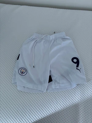 Manchester City vita träningsshorts - Säljer ett par vita träningsshorts från Manchester City med klubbmärke och nummer 9 på framsidan. Shortsen har resårmidja med dragsko och är tillverkade i ett lätt och ventilerande material. Perfekta för fotbollsträning eller gymmet.
