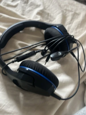 Svarta gaminghörlurar med mikrofon - Säljer ett par svarta gaminghörlurar med blå detaljer och inbyggd mikrofon. Hörlurarna har mjuka öronkuddar i tyg och justerbart huvudband. Kabeln har inbyggd kontroll för ljud och mikrofon. Perfekt för gaming eller videosamtal.