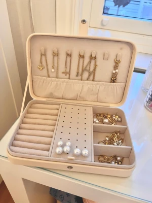 Beige smyckeskrin  - Snyggt beige smyckeskrin i läderimitation med flera fack och hållare för örhängen, ringar och halsband. Innehåller olika örhängen i guldton och pärlor samt några hängen och kedjor. Perfekt för att organisera dina favoritsmycken.