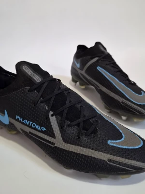 Nike Phantom ELite sg pro EU 44 - Säljer ett par Nike Phantom fotbollsskor i svart med grå och blå detaljer. Skorna har snörning och är tillverkade i syntetmaterial för bästa grepp och känsla på planen. Perfekta för dig som vill sticka ut med stilrena och sportiga skor på fotbollsplanen.