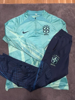 Nike Brasil träningsoverall blå/turkos - Säljer en snygg Nike Brasil träningsoverall med långärmad turkos tröja och matchande mörkblå byxor. Tröjan har halv dragkedja, grafiskt mönster och Brasil-logga på bröstet. Byxorna har dragkedja vid fickorna och samma logga. Perfekt för fotbollsträning eller chill.