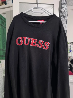 Svart stickad tröja från Guess - Snygg svart stickad tröja från Guess med stor röd och vit logga broderad på bröstet. Tröjan har rund halsringning och ribbade muddar vid ärmslut och nederkant. Perfekt för dig som gillar streetwear och vill ha en bekväm och stilren look.