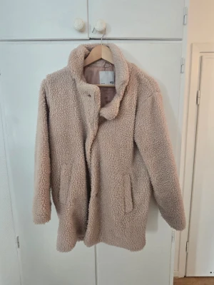 Beige teddyjacka från Lager 157 - Mysig beige teddyjacka från Lager 157 med hög krage och två fickor framtill. Jackan har en fluffig och mjuk yta i teddy-material och stängs med knappar. Perfekt för kalla dagar när du vill vara både varm och snygg.