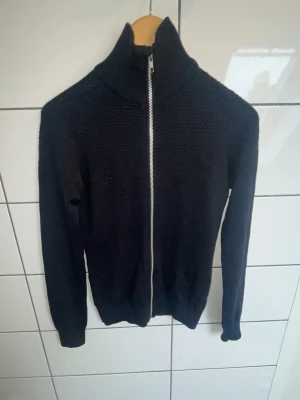 Samøe & Samøe Arno Zip - Säljer en stilren och tidlös stickad cardigan från Samsøe & Samsøe, modell Arno Zip. Tröjan är mörkblå med hög ribbstickad krage, hel dragkedja i metall och ett snyggt strukturstickat parti över bröst och rygg. Perfekt för både vardag och mer uppklädda tillfällen. Storlek: S Färg: Mörkblå/Navy Modell: Arno Zip Skick: Bra begagnat skick Nypris för liknande stickade ziptröjor från Samsøe & Samsøe ligger omkring 1 400-1 800 kr.
