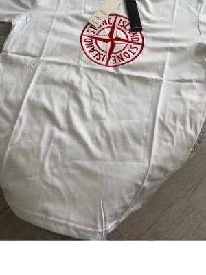 Vit t-shirt med rött Stone Island-tryck - Vit t-shirt från Stone Island med stort rött logotryck på bröstet. Klassisk passform och rund halsringning. Perfekt till jeans eller shorts för en clean och stilren look.