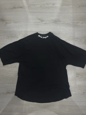 Svart Palm Angels t-shirt - Svart t-shirt från Palm Angels med stort vitt logotryck på ryggen och mindre tryck i nacken. Oversized passform och korta ärmar. Perfekt för dig som gillar streetwear och vill sticka ut med en ikonisk look.