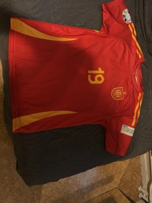 Spansk landslagströja nr 19 röd - Säljer en röd fotbollströja från Spaniens landslag med gula detaljer och nummer 19. Tröjan har trycket 'Lamine Yamal' på ryggen och landslagets emblem på bröstet. Perfekt för dig som vill stötta Spanien under matcherna!