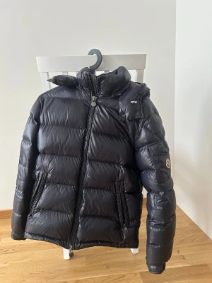  Moncler Maya mörkblå storlek 2/S - Säljer en svart, glansig dunjacka från Moncler med huva och dragkedja framtill. Jackan har två framfickor med dragkedja och en patch med Moncler-logga på ärmen. Insidan har en serietidningsdetalj och innerficka med dragkedja. Perfekt för kalla vinterdagar.