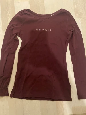 Mörkröd långärmad topp från Esprit - Snygg mörkröd långärmad topp från Esprit med rund halsringning och diskret logga i strass framtill. Tillverkad i mjukt material som sitter skönt mot kroppen. Perfekt att styla med jeans eller kjol för en clean look.