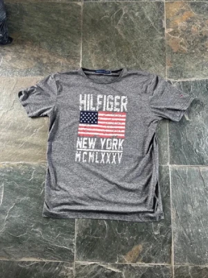 Grå Hilfiger t-shirt med USA-flagga - Snygg grå t-shirt från Hilfiger med tryck av amerikanska flaggan och texten 'NEW YORK MCMLXXXV' på bröstet. T-shirten har rund hals och korta ärmar, perfekt för en avslappnad stil.
