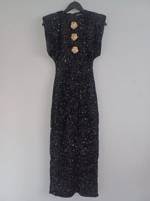 Full-length sequin evening dress - Elegant full-length sequin evening dress with exaggerated padded shoulders for a strong, flattering silhouette. Figure-hugging design with a stretchy fit, back zipper and a stylish slit at the bottom for easy movement.  Features three rose-shaped accessories at the front, which can be cut off and replaced if desired. Soft velvet-like outer fabric gives the dress a luxurious look.  Size: S / 36 Condition: New without tags. Colour: Black Material: 100% polyester  Measurements: • Length: 134 cm • S