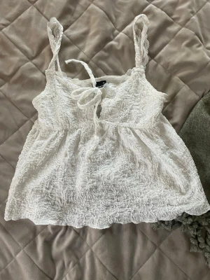 Vit linnetopp med spets från Gina Tricot - Supersöt vit linnetopp från Gina Tricot med spetsdetaljer och knyt framtill. Toppen har en skrynklig struktur och tunna axelband, perfekt för en fräsch och somrig look.