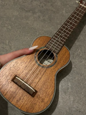 Ukulele i trä med naturfinish - Säljer en snygg ukulele i trä med naturlig finish och vita detaljer runt kroppen. Fyra strängar och klassisk form, perfekt för att jamma hemma eller med vänner. Lätt och smidig att ta med sig överallt.
