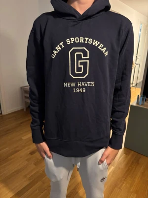 Mörkblå hoodie från Gant - Snygg mörkblå hoodie från Gant med stor logga och texten 'Gant Sportswear New Haven 1949' i vitt på bröstet. Klassisk huva och ribbade muddar vid ärmar och nederkant. Perfekt för en avslappnad och sportig stil.