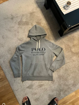Ralph Lauren Hoodie - Strlk L. Pris diskuteras privat! Säljer en grå hoodie från Polo Ralph Lauren med trycket 'POLO DENIM & SUPPLY' på bröstet. Hoodien har vita dragsnören, stor känguruficka framtill och ribbade muddar vid ärmar och nederkant. Perfekt för en avslappnad och stilren look.