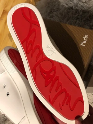 Vita sneakers med nitar från Louboutin - Snygga vita sneakers från Christian Louboutin med ikonisk röd sula och coola nitar på tån. Skorna har lågt skaft, vita skosnören och är tillverkade i slätt läder. Perfekta för dig som vill sticka ut med exklusiv design och edgy detaljer.