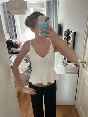 Vit peplum topp med v-ringning - Säljer en vit peplum topp med smala axelband och v-ringning fram. Toppen har en figurnära passform upptill och en volang nedtill som ger en snygg siluett. Perfekt till jeans eller kjol för en fräsch look.