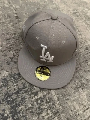 Grå LA-keps från New Era 59FIFTY - Snygg grå keps från New Era 59FIFTY med LA-logga broderad framtill i vitt. Klassisk flat peak och broderade ventilationshål. Kepsen har MLB-logga baktill och sticker ut med sin stilrena design.
