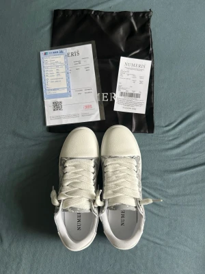 Vita sneakers från Numeris - Snygga vita sneakers från Numeris med gråa detaljer och mönstrad sida. Skorna har vita snören och en klassisk låg siluett. Ovandelen verkar vara i textil och syntet, med bekväm vadderad insida. Perfekta för en clean och stilren look.