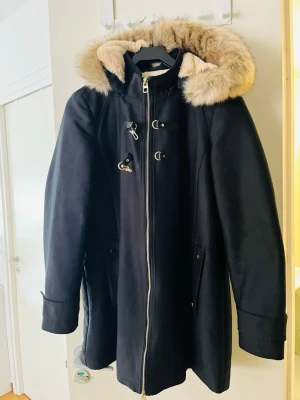 Svart vinterjacka från Mango - Snygg svart kappa från Mango med dragkedja och dekorativa spännen framtill. Kappan har en stor huva med fluffig fuskpäls i beige, perfekt för kalla dagar. Två fickor med lock på sidorna och en rak, längre passform som ger en stilren look.