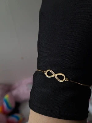 Guldigt armband med infinitysymbol - Snyggt armband i guldton med en infinitysymbol i mitten. Ena sidan av symbolen är dekorerad med små glittrande stenar för extra bling. Tunt kedjeband och justerbar låsning gör det enkelt att anpassa storleken.