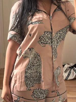 Rosa pyjamas med leopardmönster - Säljer en kortärmad pyjamas i ljusrosa med svartvita leoparder som tryck. Setet består av skjorta med knappar och bröstficka samt matchande shorts. Materialet ser ut att vara mjukt och glansigt, perfekt för chillkvällar hemma.