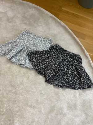 Två blommiga kortkjolar - Säljer två söta kortkjolar med blommigt mönster. Den ena är svart med vita blommor och den andra är ljusgrå med vita och svarta blommor. Kjolarna har en lätt utsvängd form! 
