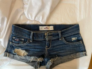 Mörkblå jeansshorts från Hollister - Snygga mörkblå jeansshorts från Hollister med slitna detaljer och upprullade benslut. Klassisk femficksmodell med dubbla knappar framtill och broderad logga på fickan. Perfekta för varma dagar och en avslappnad stil.