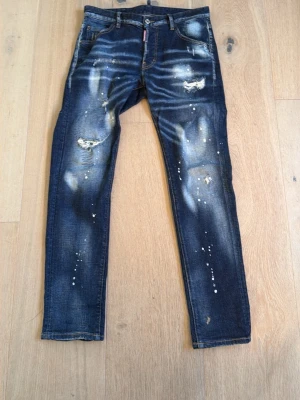 Mörkblå dsquared jeans - Säljer ett par mörkblå jeans från Ciao Bella med slitningar, hål och vita färgstänk för en riktigt edgy look. Jeansen har smal passform och coola detaljer både fram och bak. Perfekta för dig som gillar streetstyle och vill sticka ut.