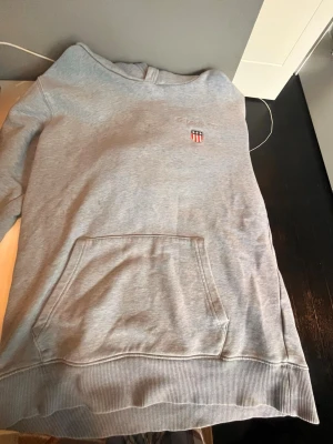 Ljusgrå hoodie med magficka - Snygg ljusgrå hoodie med huva och stor magficka framtill. På bröstet finns ett litet broderat märke med sköld och text. Hoodien har ribbade muddar vid ärmslut och nederkant. Perfekt för en chill och avslappnad stil.