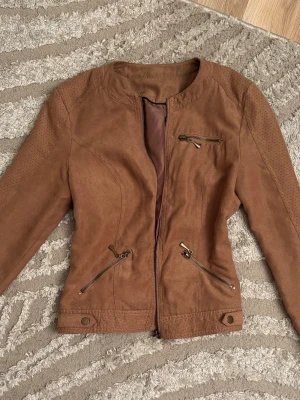 Brun skinnjacka med dragkedjor - Snygg brun skinnjacka i bikerstil med flera dragkedjor framtill och på ärmarna. Jackan har rundad krage, dekorativa sömmar och detaljerade axelpartier. Perfekt för dig som vill ha en stilren och trendig look.
