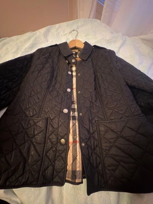 Svart quiltad jacka med rutigt foder - Snygg svart quiltad jacka med stora framfickor och klassisk krage. Insidan har ett rutigt foder i beige, rött och grått. Jackan stängs med knappar framtill och har en rak passform som passar till många olika stilar.