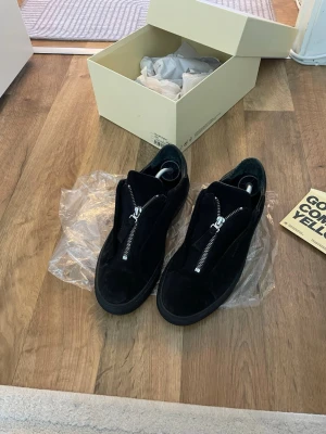 Svarta sneakers med dragkedja från Axel Arigato  - Säljer ett par svarta sneakers i mocka med cool dragkedja framtill. Skorna har en låg silhuett och svart sula, vilket ger en stilren och modern look. Perfekta för dig som gillar minimalistisk design och vill sticka ut lite extra. Storlek 41 