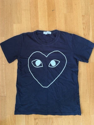 Marinblå  Comme des Garçons Play t-shirt - Säljer en marinblå t-shirt från Comme des Garçons Play med det ikoniska hjärtmotivet med ögon på bröstet. T-shirten har rund hals och korta ärmar. Perfekt för dig som gillar streetwear och vill sticka ut med en unik design.  Passar s/xs 