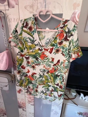 Blommig topp från H&M, storlek 36 - Säljer en färgglad blommig topp från H&M i storlek 36. Toppen har korta ärmar, v-ringning och ett tropiskt mönster med blommor och fjärilar i grönt, rött och orange på vit botten. Perfekt för vår och sommar!