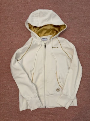 Vit hoodie från Reebok Classic - Snygg vit hoodie från Reebok Classic med dragkedja och guldfärgat innerfoder i huvan. Har gulddetaljer vid sömmarna och en liten broderad logga vid fickan. Perfekt för dig som vill ha en stilren och sportig look.