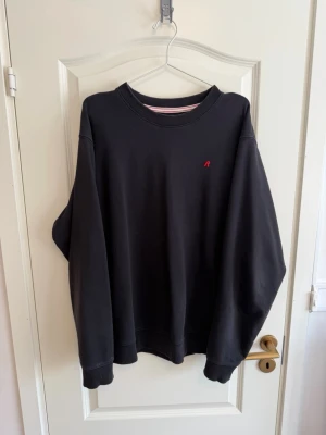 Svart sweatshirt med röd detalj - En stilren svart sweatshirt med rund halsringning och en liten röd broderad bokstav på bröstet. Tröjan har ribbade muddar vid ärmslut och nederkant. Perfekt för en avslappnad och trendig look.