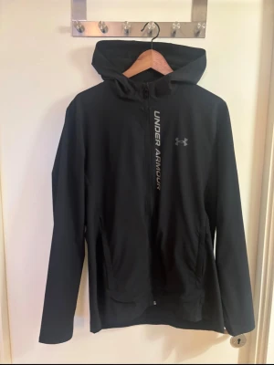 Under Armour LöparJacka - Svart träningsjacka från Under Armour i storlek M. Stilren och lätt modell med huva, dragkedja framtill och fickor. Under Armour-logga samt logotypdetaljer framtill och reflexdetalj baktill.  Jackan är i fint begagnat skick, se bilder för skick.  • Storlek: M • Färg: Svart • Under Armour • Huva • Dragkedja framtill • Sidofickor • Kvitto finns ej