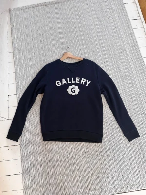 Mos mosh tröja  - Mörkblå sweatshirt med vitt GALLERY-tryck och en broderad blomma med bokstaven G på bröstet. Tröjan har rund hals, långa ärmar och ribbade muddar. Liten patch med G på ärmslutet. Perfekt för en avslappnad och trendig look.