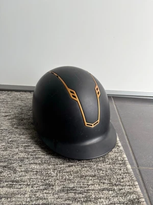 Svart ridhjälm med gulddetaljer - Snygg svart ridhjälm med mattsvart finish och coola guldfärgade detaljer framtill. Hjälmen har en klassisk rundad form och liten skärm framtill. Perfekt för dig som vill ha en stilren och modern look i stallet. Ridhjälmens storlek finns med i bilderna
